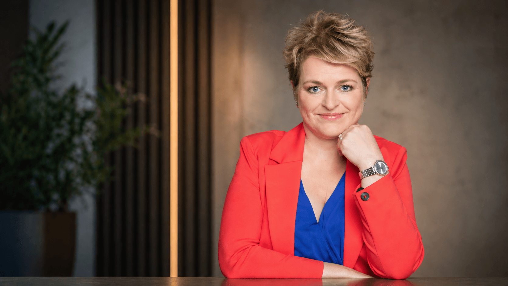 Professional woman with short blonde hair in a red blazer and blue blouse, resting her chin on her hand at a desk.