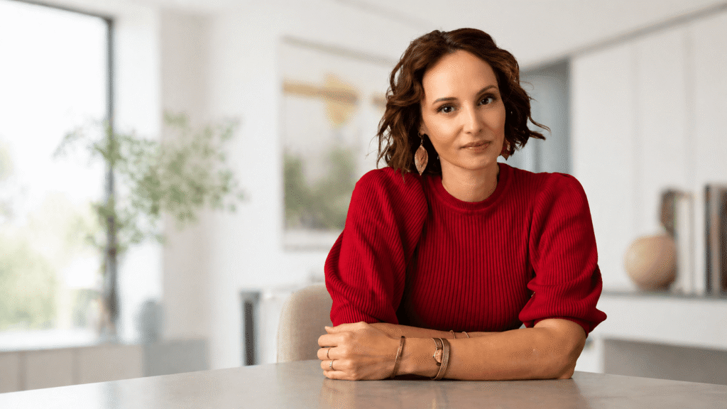Woman with short brown hair in a red ribbed sweater sits at a light table in a bright, modern room, looking at the camera with a gentle smile.