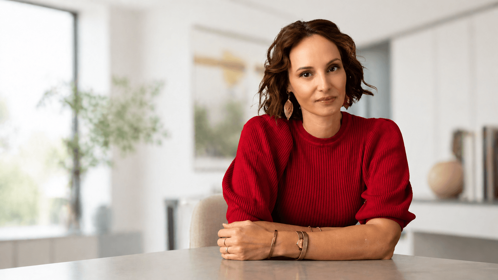 Woman with short brown hair in a red ribbed sweater sits at a light table in a bright, modern room, looking at the camera with a gentle smile.